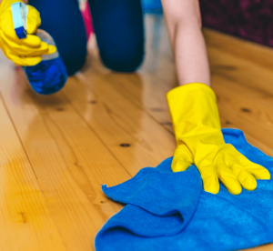 person wearing yellow rubber gloves, kneeling and cleaning a hardwood floor with a blue microfibre cloth